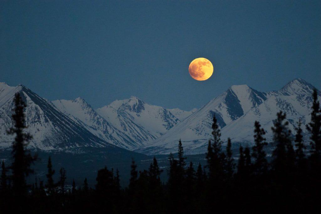 A full moon rises over a snowy Alaska mountain range.