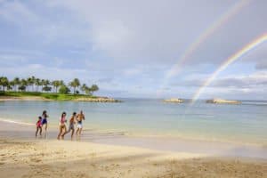 Three people standing on a beach at Aulani Disney Resort with a rainbow in the sky.