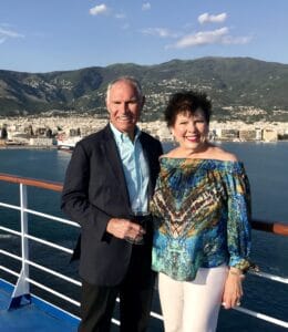 An older couple smiling on a Fox Travel cruise ship deck with a coastal city and mountains in the background.