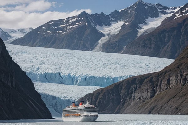 A cruise ship from Fox Travel approaches a large glacier between mountains under a partly cloudy sky.