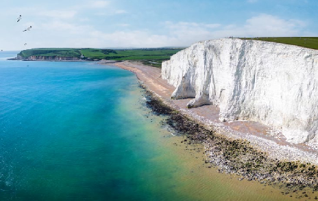 Aerial view of the White Cliffs of Dover with a turquoise sea and clear sky, seagulls flying, and a pebbly beach extending into the distance; imagine witnessing a solar eclipse on one of those serene cruises.