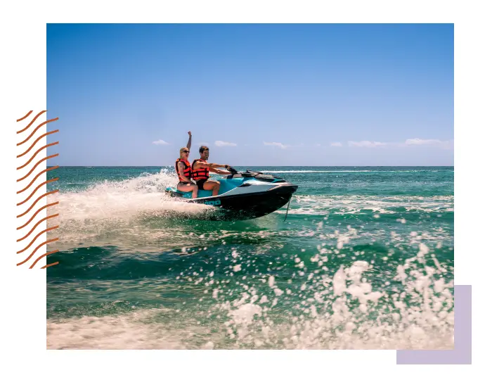 Two people wearing life jackets riding a jet ski on a sunny day, creating a splash in the turquoise water. MORE INCLUSIVE™, their joy radiates in every wave and ripple.