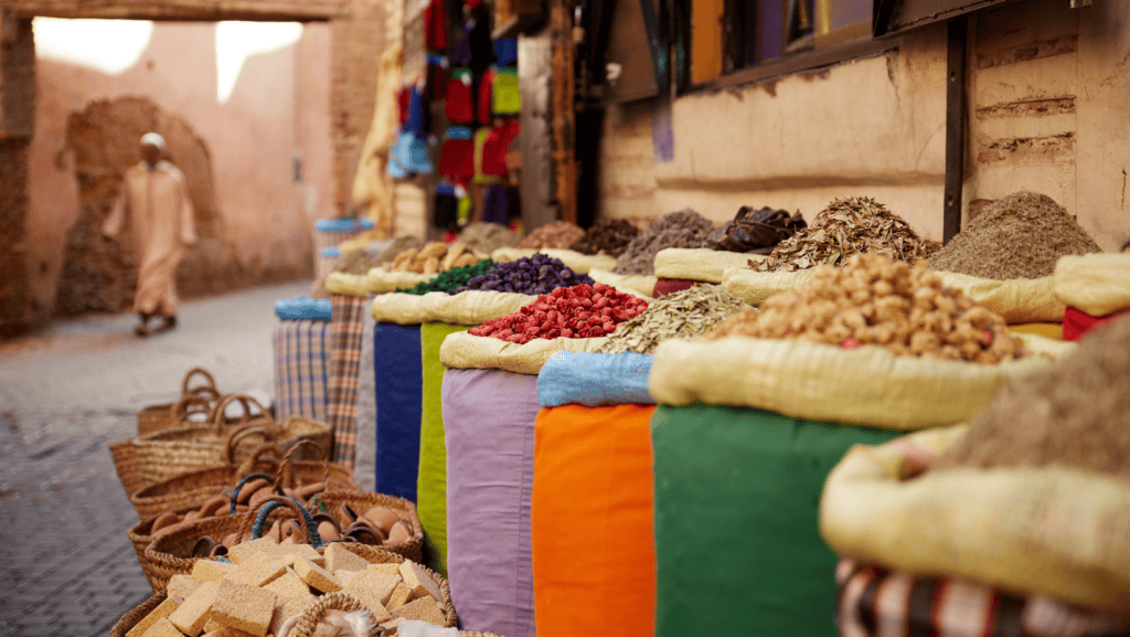 Bags of colorful spices and nuts are displayed along a street with woven baskets, evoking the charm of exotic markets. A person walks in the background, wearing traditional attire, adding to the allure of this scene befitting romantic getaways.