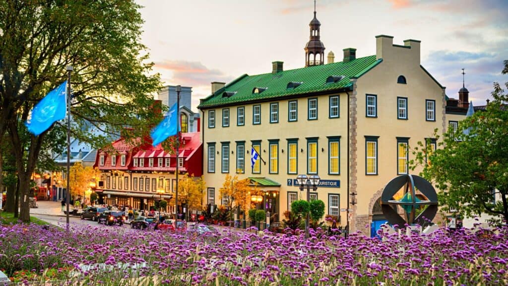 Street view of a charming European-style building with a green roof and colorful shutters, ideal for romantic getaways. It's surrounded by purple flowers and trees, all under a sunset sky.