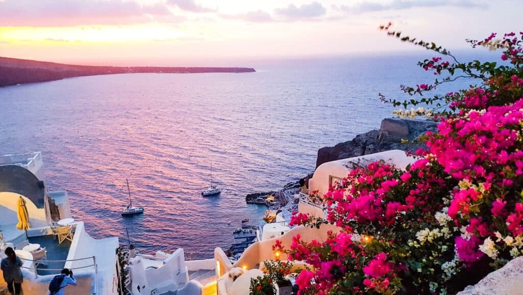 A romantic getaway awaits with a sunset view over a coastal town, where white buildings contrast with the vibrant pink bougainvillea and sailboats drift peacefully on the shimmering water.