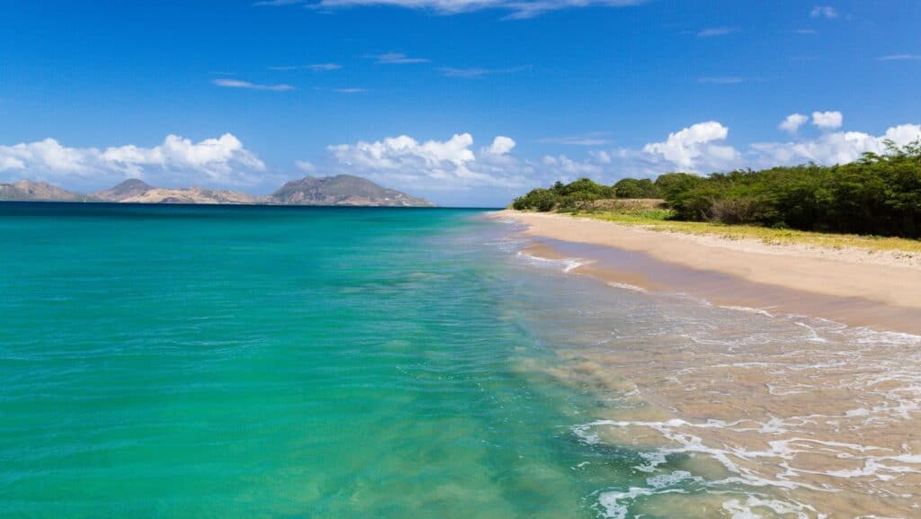 A serene beach with turquoise water, gentle waves, and a sandy shore—perfect for romantic getaways. In the distance, green hills rise under a partly cloudy blue sky.