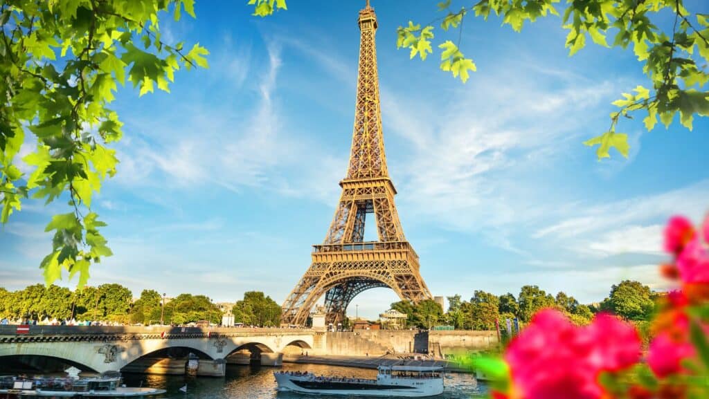 The Eiffel Tower stands tall against a blue sky, framed by green leaves and pink flowers—a perfect backdrop for romantic getaways. Below, boats gently cruise along the Seine River.