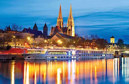 A Danube river cruise ship is docked by a European riverside, its silhouette framed against the illuminated buildings and twin-spired cathedral reflecting in the water at dusk—a perfect scene curated by Travel Agency The Woodlands.