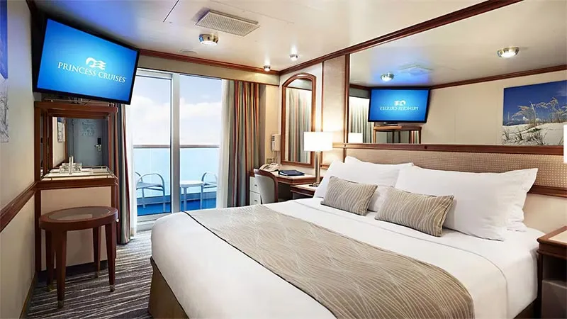 Experience a spacious Majestic Princess cruise ship cabin featuring a large bed, two TVs, a desk, and a balcony with chairs—perfect for relaxing during your British Isles cruise with Fox Travel. Neutral decor adds to the comfort.