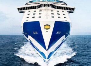A large cruise ship with a blue and white hull sails forward on the open sea, creating waves at the bow—perfect for those seeking exclusive travel promotions from American Express Travel.