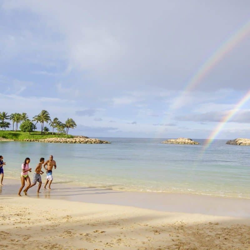 Three people standing on a beach at Aulani Disney Resort with a rainbow in the sky.