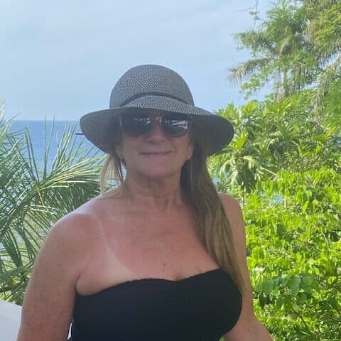 A woman in sunglasses and a sun hat stands on a balcony with tropical greenery and the ocean in the background, wearing a black strapless top, proudly displaying her Team Fox logo.